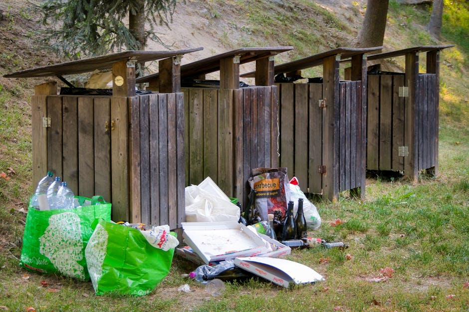 Four wooden outdoor rubbish bins with slanted, slightly weathered dark brown lids are positioned in a row on a grassy area, with the containers made of vertical wooden planks. In front of the bins, th