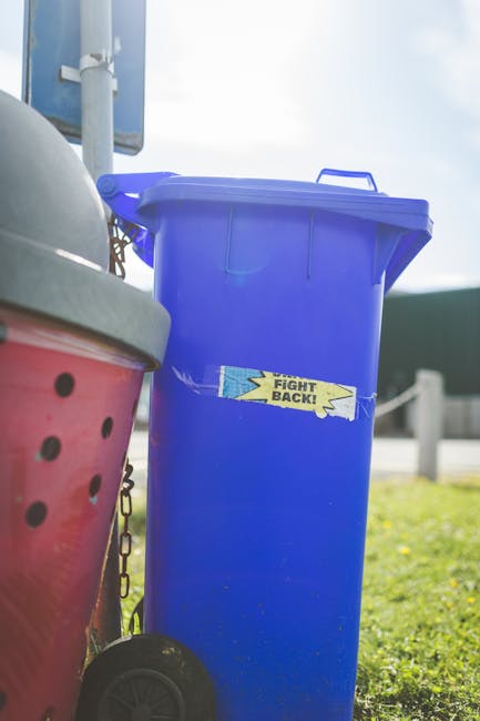 Four wooden outdoor rubbish bins with slanted, slightly weathered dark brown lids are positioned in a row on a grassy area, with the containers made of vertical wooden planks. In front of the bins, th