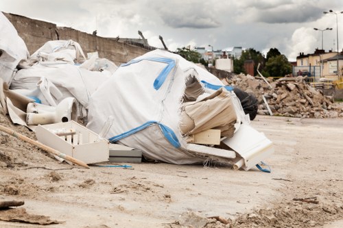 A large pile of construction and renovation debris, including white plastic bags filled with rubbish, insulation panels, and various broken building materials, is situated on a dirt surface outdoors. The debris appears to include items such as foam insulation, PVC pipes, and cardboard or fiberboard pieces, with some bags tied or partially open, revealing contents inside. In the background, there is a concrete wall with some exposed rebar, and further back, a row of trees and residential or commercial buildings can be seen under a cloudy sky. The scene suggests an outdoor rubbish removal or clearance activity, typical of construction site cleanup or garden clearance services in Bermondsey, as handled by Garden Clearance Bermondsey. The ground surface is dry and uneven, with loose soil, and the entire setting conveys a sense of large-scale waste collection in the process of being sorted or prepared for disposal, aligning with professional rubbish removal practices in the local area.