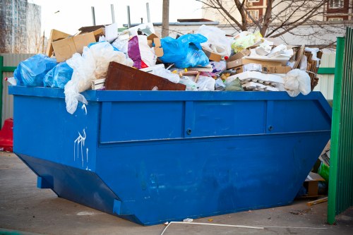A large blue metal skip positioned on a paved surface outdoors, filled with assorted waste materials including cardboard boxes, white plastic bags, torn paper, and pieces of fabric. The skip is situated near a green metal fence with vertical bars on the right side, and there are leafless trees and residential buildings visible in the background, suggesting a borough location such as Bermondsey. The waste occupies the entire volume of the skip, with some items slightly overflowing the top edges, and the surface of the waste displays varied textures and colors, predominantly brown, white, and blue. The overall scene captures a typical rubbish removal setup for garden clearance or building waste disposal, managed by Garden Clearance Bermondsey, with clear evidence of a professional rubbish collection environment intended for local waste management services.