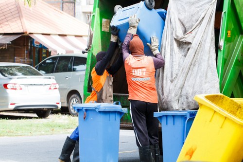 Two waste collection workers wearing orange high-visibility vests and protective gloves are shown lifting a large blue plastic barrel into the back of a green rubbish collection truck in a residential area. The truck has a grey tarpaulin partially covering its loading area. In the foreground, there are two additional blue bins and a yellow recycling container on the street pavement. Behind the scene, parked cars and a house with a brown and white striped awning are visible, indicating a typical Bermondsey street environment. The workers are focused on their task, highlighting the practical aspects of rubbish removal services provided by Garden Clearance Bermondsey, which caters to local garden and general waste clearance needs in the borough. The overall atmosphere is functional, with natural daylight illuminating the scene, emphasizing the process of efficient rubbish collection in an urban setting.